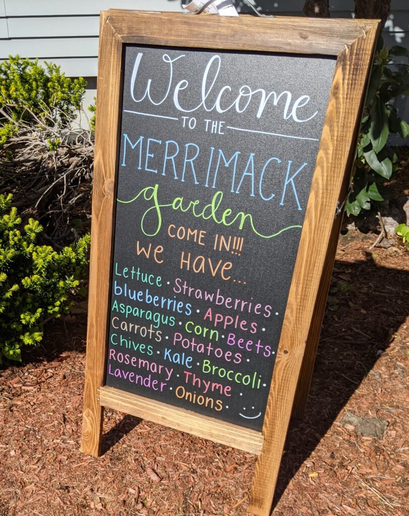 A chalk board sign welcomes people to the Merrimack Garden. We have lettuce, strawberries, blueberries, apples, asparagus, corn, beets, carrots, potatoes, chives, kale, broccoli, rosemary, thyme, lavender, onions. 