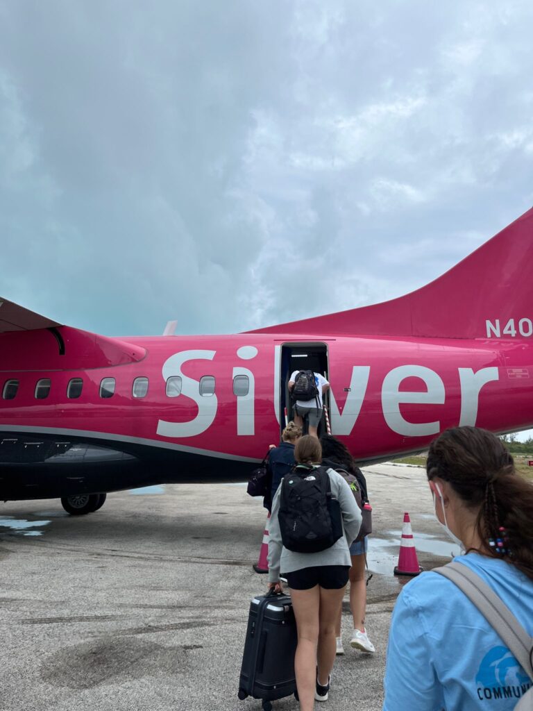 Alt text: People carrying luggage walk in a single file line into the left rear door of a small pink airplane. 
