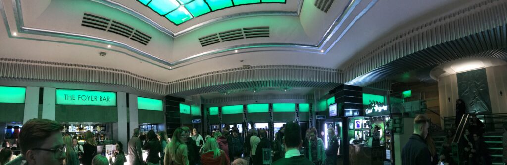 A panoramic image of a busy theatre lobby decorated in green lights. (Photo credits: Emma McDonald, 2017)