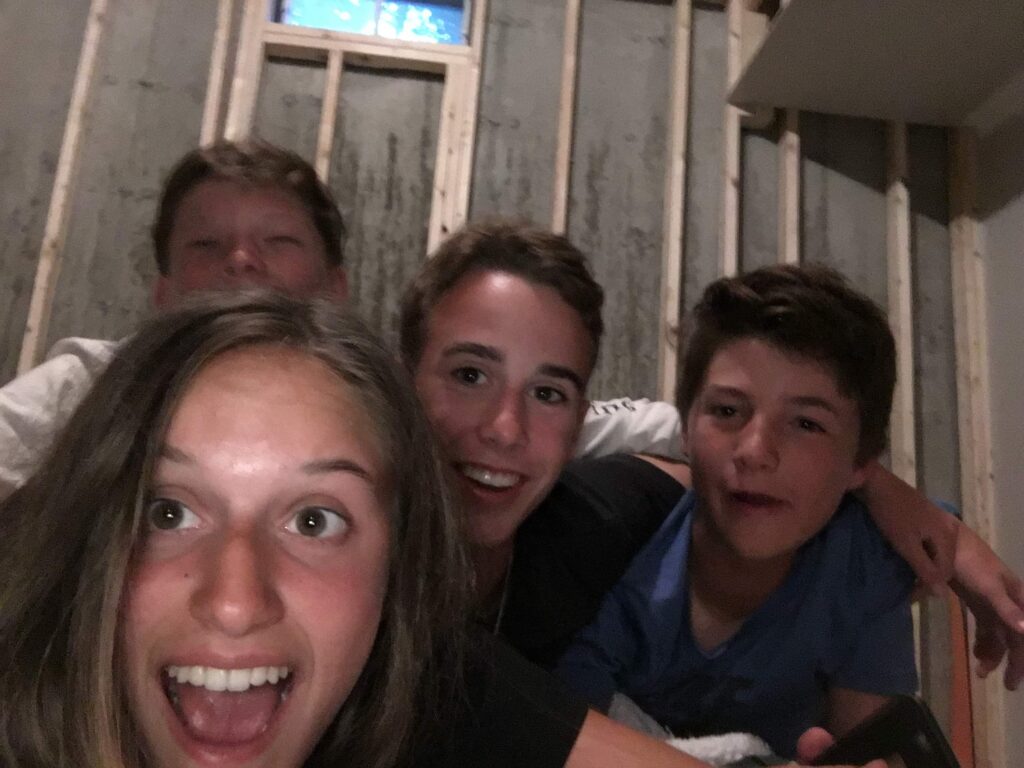 Four young teens in a basement taking a selfie—three boys and one girl (the girl in front holding the phone), all smiling and crowded together.