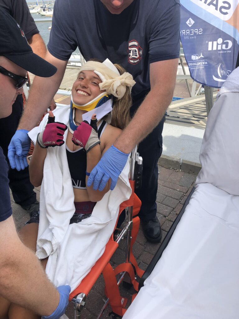 Katie being wheeled from the dock in a wheelchair, holding 2 thumps up with bandages around her head, a neck brace, pink sailing gloves, and an orange safety belt, surrounded by paramedics assisting her.