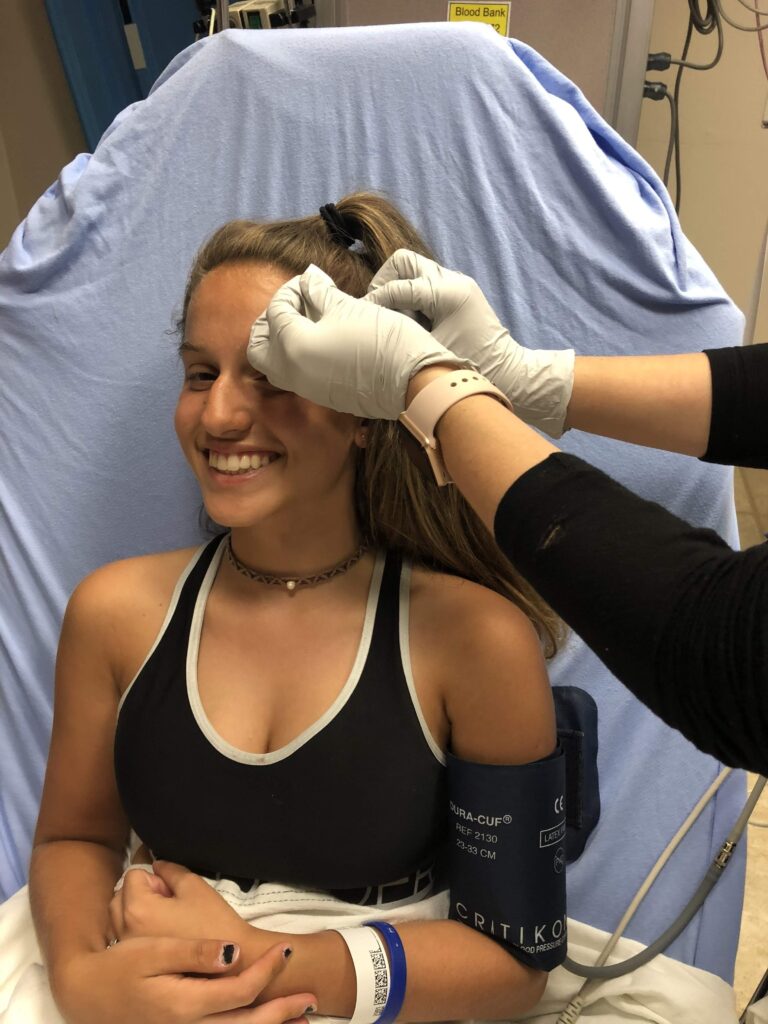 Katie lying on a hospital bed with blue sheets in the emergency room, still smiling while getting stitches in her head, with a blood pressure cuff on her arm and a white hospital blanket covering her lower half.