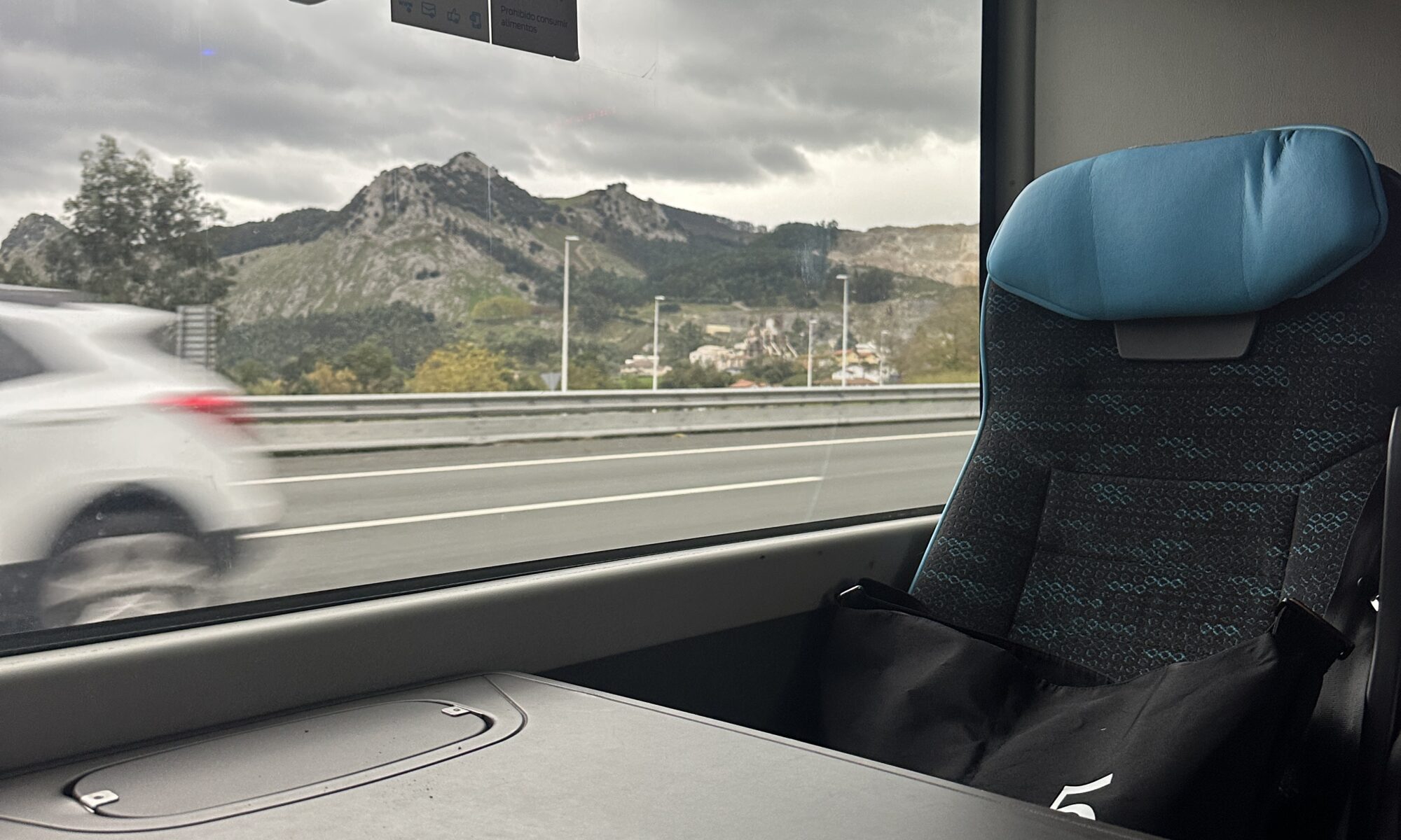 Bus table and seat. Window with landscape in the background. Sky is cloudy.