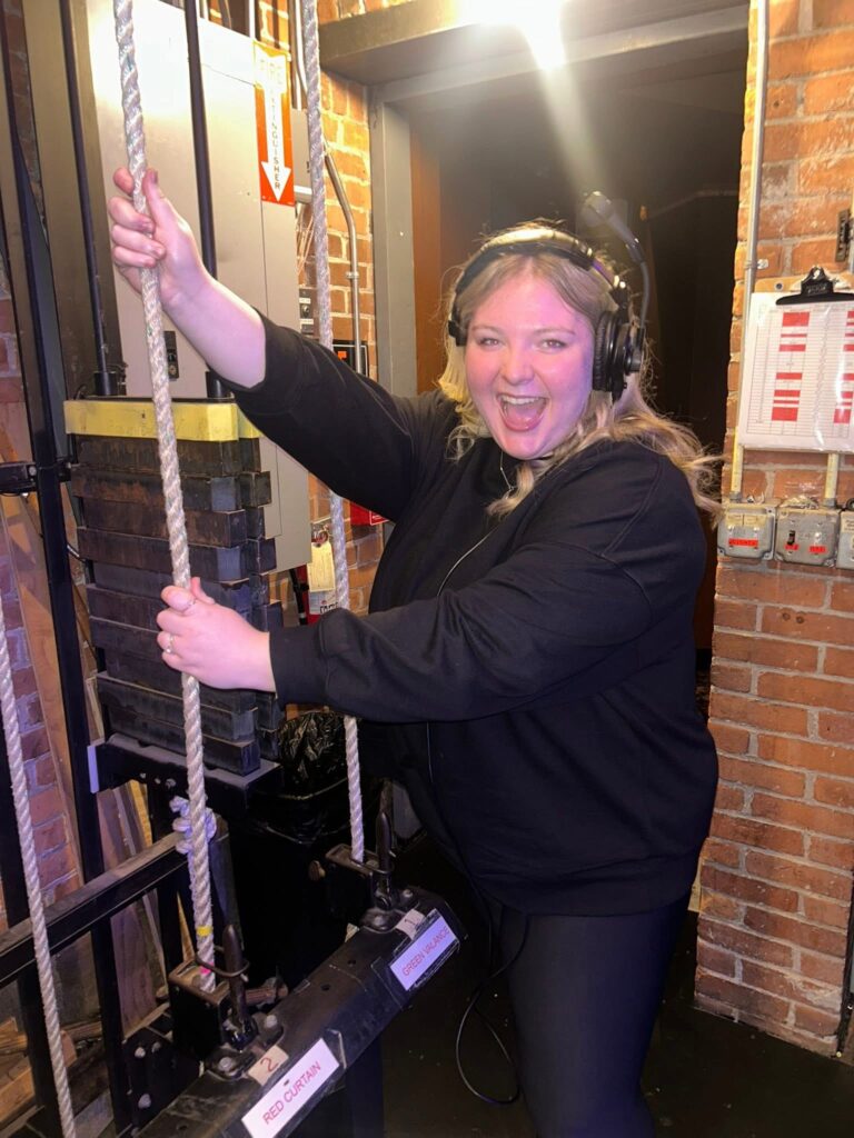 A smiling young woman wearing all black clothing stands in front of a brick wall holding the curtain rope backstage at a theatre.