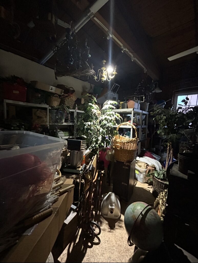 A medium sized room filled with shelves full of prop bins used for theatrical performance. A vintage wheelchair, a houseplant, some wicker baskets. There is a single-bulb light on the exposed ceiling.