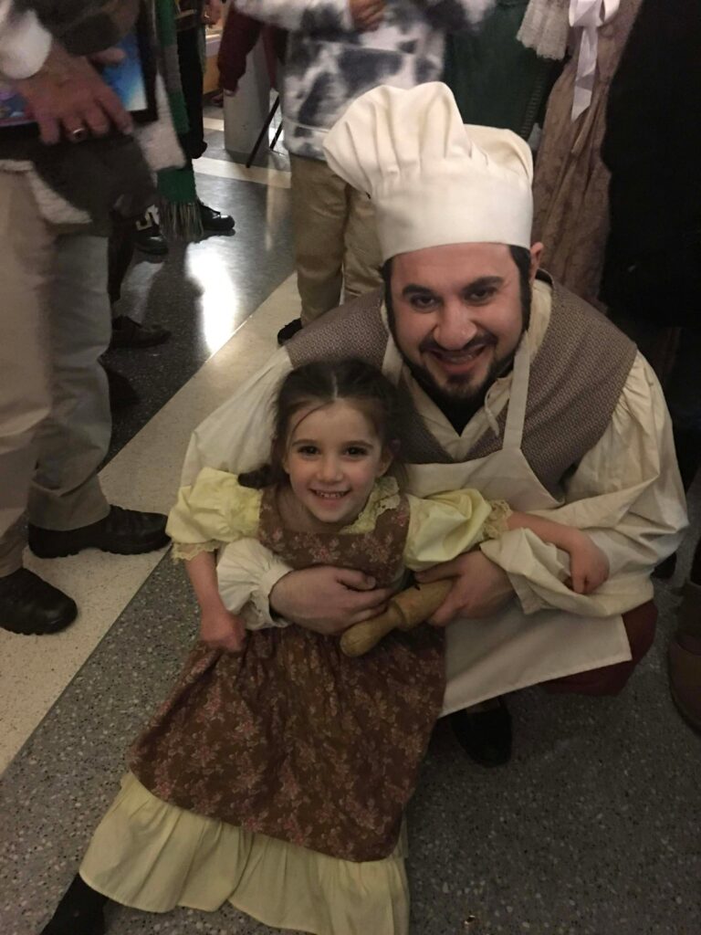 A man hugs his young daughter as both pose and smile. They are dressed up in simple French peasant costumes, reminiscent of a Disney film.